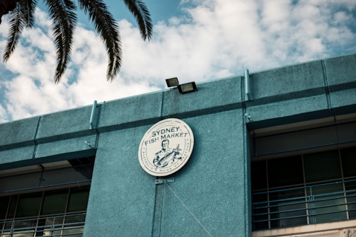 A circular sign for the Sydney Fish Market is prominently displayed on the exterior of a blue building. The sign features a graphic and writing, with palm leaves partially visible above. The sky in the background is filled with scattered clouds.