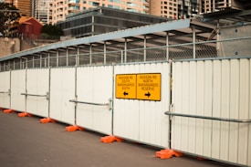 A construction site with tall metal barricades featuring directional signs for Hickson Road South and North Barangaroo Reserve. The path is lined with orange barricades and modern office buildings are visible in the background.