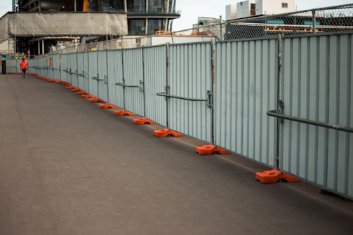 A long, grey metal fence lines the right side of a deserted paved pathway. The fence is supported by orange bases. In the distance, there is a person wearing an orange vest, possibly a construction worker. Behind the fence, buildings in various stages of construction are visible, with scaffolding and construction materials present.