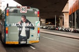A street scene featuring the rear view of a city bus displaying an advertisement for a clothing brand. The bus is situated on a road lined with numerous parked motorcycles under a modern architectural structure. The advertisement features a person wearing a jacket with the brand's name. Traffic and informational signs are visible on the bus's rear.