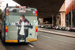 A street scene featuring the rear view of a city bus displaying an advertisement for a clothing brand. The bus is situated on a road lined with numerous parked motorcycles under a modern architectural structure. The advertisement features a person wearing a jacket with the brand's name. Traffic and informational signs are visible on the bus's rear.