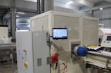 A large industrial machine with a control panel and a digital touchscreen interface mounted on it. The environment is a factory setting with visible overhead lights and other machinery in the background. There are various buttons and emergency stop features on the equipment, along with visible wiring and mechanical parts.