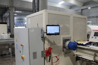 A large industrial machine with a control panel and a digital touchscreen interface mounted on it. The environment is a factory setting with visible overhead lights and other machinery in the background. There are various buttons and emergency stop features on the equipment, along with visible wiring and mechanical parts.
