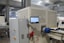 A large industrial machine with a control panel and a digital touchscreen interface mounted on it. The environment is a factory setting with visible overhead lights and other machinery in the background. There are various buttons and emergency stop features on the equipment, along with visible wiring and mechanical parts.