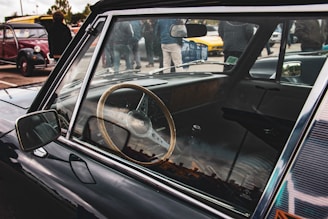 A classic car with a wooden steering wheel is parked, with a view of the dashboard visible through the window. Several people are gathered in the background, and other vintage cars are parked nearby.
