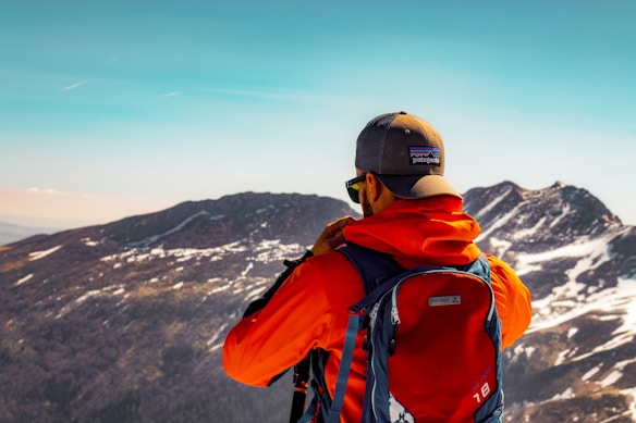 A hiker dressed in a bright orange jacket and wearing a cap is seen from behind, standing on a mountain landscape with snow-dusted peaks and a clear blue sky. He carries a large blue backpack with a visible brand logo.