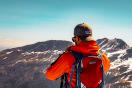 A hiker dressed in a bright orange jacket and wearing a cap is seen from behind, standing on a mountain landscape with snow-dusted peaks and a clear blue sky. He carries a large blue backpack with a visible brand logo.