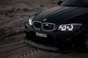 The front section of a black sports car is parked on a sandy terrain. The car has a sleek design with gleaming, illuminated headlights featuring angel eyes. The grille showcases the iconic BMW logo and distinctive M series coloring. A visible license plate with registration KL 43 D 2087 is attached to the front.