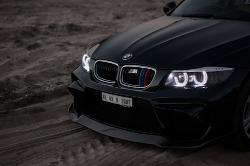 The front section of a black sports car is parked on a sandy terrain. The car has a sleek design with gleaming, illuminated headlights featuring angel eyes. The grille showcases the iconic BMW logo and distinctive M series coloring. A visible license plate with registration KL 43 D 2087 is attached to the front.