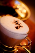 A glass cup filled with a frothy cappuccino topped with a sprinkle of cocoa powder sits on a golden tray. Warm lighting creates a cozy ambience with soft bokeh effects in the background.
