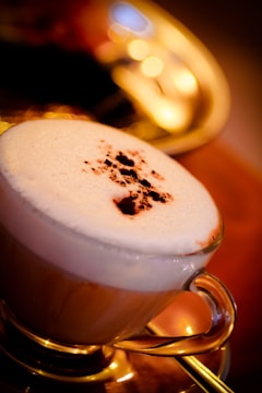 A glass cup filled with a frothy cappuccino topped with a sprinkle of cocoa powder sits on a golden tray. Warm lighting creates a cozy ambience with soft bokeh effects in the background.