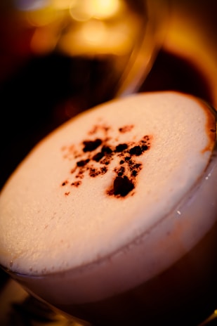 A close-up view of a drink with a frothy cream topping, sprinkled with cocoa or chocolate powder. The background is softly blurred with warm, golden bokeh lights.
