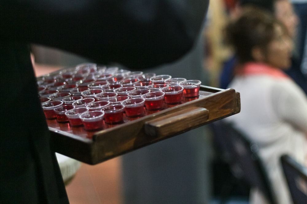 person carrying red liquid filled mini disposable cups on wooden tray