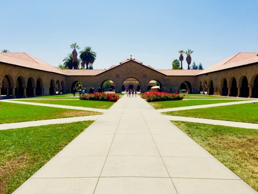 A symmetrical courtyard with a long pathway leading to an arched building. Manicured lawns and flower beds flank the pathway, while tall palm trees are visible in the background. The architecture features stone arches and red-tiled roofs, typical of a university or historical building.