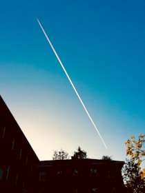 A clear blue sky is intersected by a single contrail diagonally across the scene. Below the sky, silhouettes of buildings and trees are visible, with sunlight casting a warm glow on their tops.