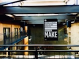 An industrial-style interior space with concrete walls and metal beams. A person walks along a metal walkway. A prominent black and white sign hangs from the ceiling, displaying motivational text. Natural light floods through skylights, casting shadows.