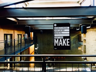 An industrial-style interior space with concrete walls and metal beams. A person walks along a metal walkway. A prominent black and white sign hangs from the ceiling, displaying motivational text. Natural light floods through skylights, casting shadows.