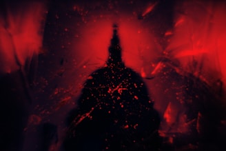 A dark, silhouetted structure reminiscent of a spire or tower is central against a vivid red backdrop. Splattered red paint and patterns suggest a chaotic or abstract artistic style.
