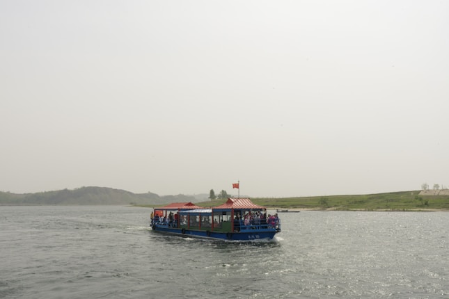 A blue and white boat with a red canopy sails across a wide river. The boat carries passengers who are observing the surrounding scenery. In the background, there are rolling green hills and a cloudy sky.