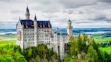 A grand, fairy-tale-like castle stands majestically on a rocky hill surrounded by lush green forests and a vibrant landscape. The castle features numerous turrets and spires, with a stunning architectural design reminiscent of medieval times. Rolling hills and distant alpine meadows span across the background under a cloudy sky.