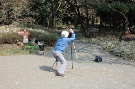 A person is intently looking through a spotting scope mounted on a tripod, seated on a path surrounded by trees and bushes. There is a sign indicating directions to the Shinjuku Gate, and a bag or case is on the ground nearby. Another person can be seen taking a photograph in the background.