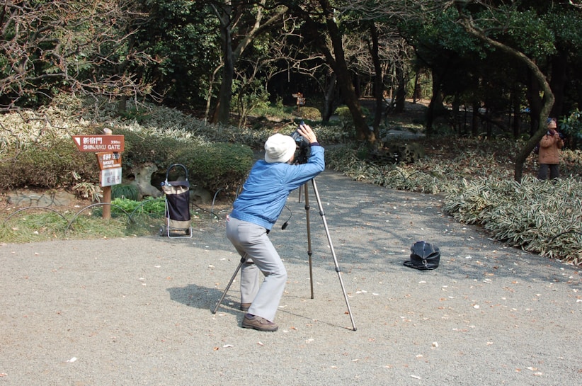 A person is intently looking through a spotting scope mounted on a tripod, seated on a path surrounded by trees and bushes. There is a sign indicating directions to the Shinjuku Gate, and a bag or case is on the ground nearby. Another person can be seen taking a photograph in the background.