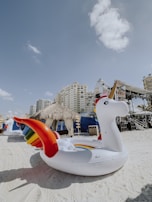 A large inflatable unicorn float with rainbow wings sits on a sandy beach. Tall buildings are visible in the background, along with a thatched-roof hut and a stage setup with scaffolding. The sky is predominantly clear with a few clouds.