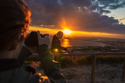 man taking photo of man beside railing under gray and orange cloudy sky with sun