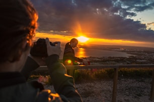 man taking photo of man beside railing under gray and orange cloudy sky with sun