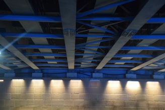 A structure with a series of blue steel beams and supports forms the ceiling, casting a geometric pattern. Below, a stone or concrete wall is illuminated by a row of evenly spaced lights, creating a contrast between the cool blue tones of the beams and the warm light on the wall.