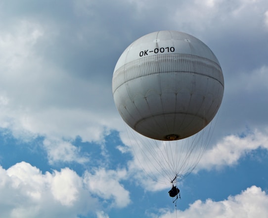 A large white spherical balloon hovers against a partly cloudy sky. The balloon appears to have a small basket or equipment suspended beneath it, secured with multiple cables. The sky is a mix of blue with scattered white clouds.