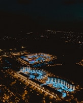 Aerial view of a brightly lit resort at night with numerous pools illuminated in blue. The complex is surrounded by a dark landscape, and the lights create a vivid contrast against the night sky.