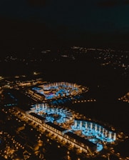 Aerial view of a brightly lit resort at night with numerous pools illuminated in blue. The complex is surrounded by a dark landscape, and the lights create a vivid contrast against the night sky.