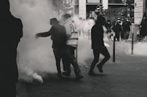 Several people are standing and moving through a smoky environment on a city street, suggesting a tense or chaotic situation. Visibility is obscured by the smoke, and there are faint silhouettes in the background.