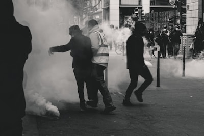 Several people are standing and moving through a smoky environment on a city street, suggesting a tense or chaotic situation. Visibility is obscured by the smoke, and there are faint silhouettes in the background.