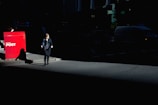 A woman walks on a dimly-lit urban street, her silhouette partially illuminated by a beam of sunlight against a backdrop of shadows. Nearby, a bright red Australia Post mailbox stands out sharply in contrast against the dark surroundings.