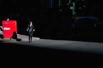A woman walks on a dimly-lit urban street, her silhouette partially illuminated by a beam of sunlight against a backdrop of shadows. Nearby, a bright red Australia Post mailbox stands out sharply in contrast against the dark surroundings.