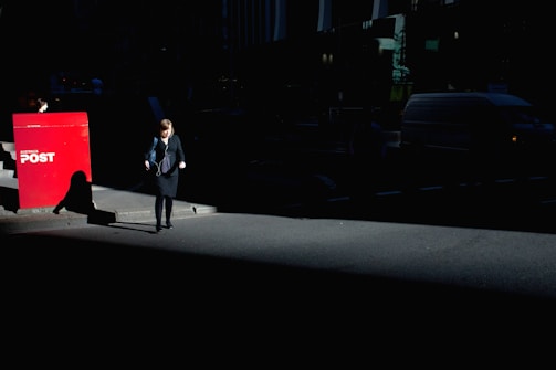 A woman walks on a dimly-lit urban street, her silhouette partially illuminated by a beam of sunlight against a backdrop of shadows. Nearby, a bright red Australia Post mailbox stands out sharply in contrast against the dark surroundings.
