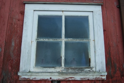 A rustic wooden window with four frosted glass panes set in a weathered red wooden wall. The paint on the white window frame is chipped and peeling, giving a vintage and aged appearance.