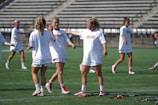 Several female athletes are on a lacrosse field, wearing white uniforms with red accents. They appear to be interacting and preparing for a game. In the background, there are empty stadium bleachers. Lacrosse sticks are lying on the ground near the players.
