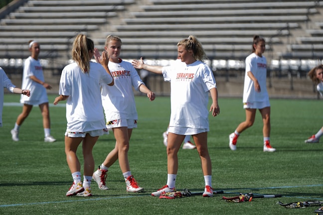 Several female athletes are on a lacrosse field, wearing white uniforms with red accents. They appear to be interacting and preparing for a game. In the background, there are empty stadium bleachers. Lacrosse sticks are lying on the ground near the players.