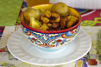 A colorful ceramic bowl filled with roasted potatoes, fried plantains, and lemon wedges sits on a white plate. The bowl features vibrant floral and geometric patterns with a color palette of blue, red, yellow, and green. It is placed on a patterned tablecloth with an assortment of colors and designs.