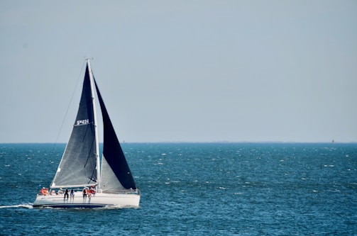 A sailboat with a dark sail is moving across a vast expanse of blue water. Several people are on the deck wearing life jackets. The sky is clear, with a pale blue hue blending into the horizon.