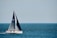 A sailboat with a dark sail is moving across a vast expanse of blue water. Several people are on the deck wearing life jackets. The sky is clear, with a pale blue hue blending into the horizon.