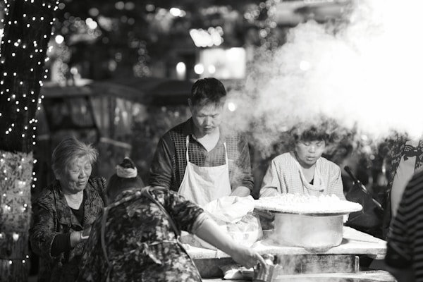 A busy street food stall where several people are engaged in cooking. Steam rises prominently from a large pot on the table, and the scene is set against a backdrop of blurred lights strung around a tree. The atmosphere feels bustling and lively, with different people focused on food preparation.