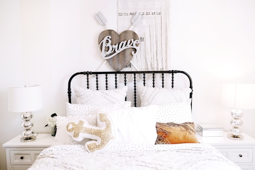 A cozy and neatly arranged bedroom with a black metal bed frame and several white pillows and blankets. Above the bed hangs a heart-shaped wooden wall decor with the word 'Brave' on it. On either side of the bed are white nightstands, each with identical metallic table lamps. A decorative anchor-shaped pillow is seen on the bed among others.