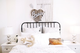 A cozy and neatly arranged bedroom with a black metal bed frame and several white pillows and blankets. Above the bed hangs a heart-shaped wooden wall decor with the word 'Brave' on it. On either side of the bed are white nightstands, each with identical metallic table lamps. A decorative anchor-shaped pillow is seen on the bed among others.