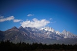 Majestic snow-capped mountains under a clear blue sky with scattered clouds. Dense green forests cover the lower slopes, creating a natural and serene landscape.
