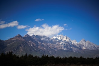 Majestic snow-capped mountains under a clear blue sky with scattered clouds. Dense green forests cover the lower slopes, creating a natural and serene landscape.