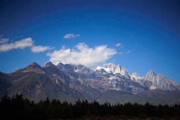 Majestic snow-capped mountains under a clear blue sky with scattered clouds. Dense green forests cover the lower slopes, creating a natural and serene landscape.